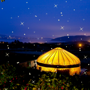 A photograph of Mulroy yurt at night time - it is lit from within so is a bright yellow colour and in the background the sea and Knockalla mountain can be seen. The picture has been overlaid with white stars